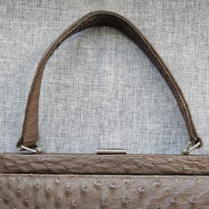 May include: A brown handbag featuring a textured ostrich-style leather finish with a single arched handle. The bag includes a rectangular metal frame closure at the top with silver-toned hardware attachments connecting the handle to the main body.