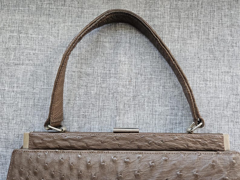 May include: A brown handbag featuring a textured ostrich-style leather finish with a single arched handle. The bag includes a rectangular metal frame closure at the top with silver-toned hardware attachments connecting the handle to the main body.