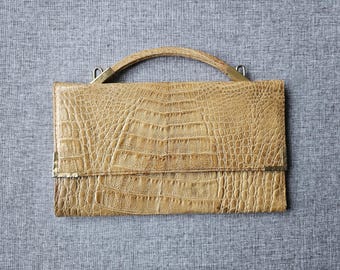 May include: A tan handbag with a textured crocodile skin pattern, featuring a curved top handle and metallic gold-toned hardware accents on the corners and handle attachments. The rectangular bag has a flap closure and a structured design.
