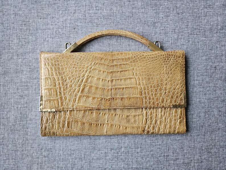 May include: A tan-coloured handbag with a crocodile skin effect. The rectangular bag features a curved handle and metal detailing. The bag is closed with a flap and is set against a grey background.