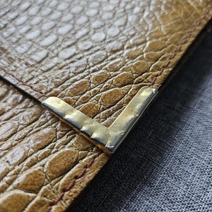 May include: Close-up of a brown leather wallet with a crocodile skin texture. The wallet has a gold-coloured metal corner protector. The stitching is visible along the edge of the wallet.