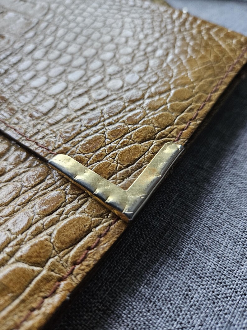 May include: Close-up of a brown leather wallet with a crocodile skin texture. The wallet has a gold-coloured metal corner protector. The stitching is visible along the edge of the wallet.