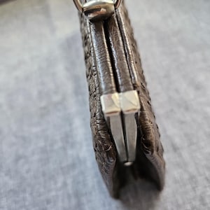 May include: A dark brown textured leather coin purse featuring a silver-toned metal kiss-lock frame closure. The exterior surface has a distinct pebbled or embossed pattern. A silver-toned metal loop is attached to the top for a strap or chain.