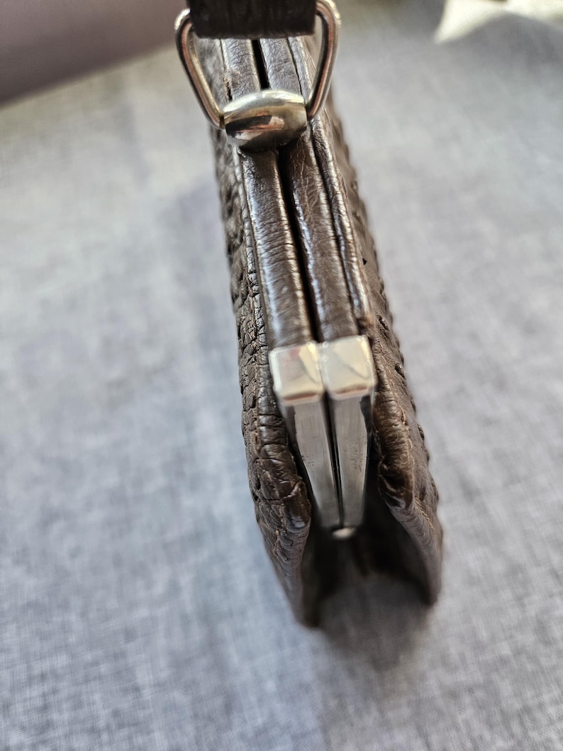 May include: A dark brown textured leather coin purse featuring a silver-toned metal kiss-lock frame closure. The exterior surface has a distinct pebbled or embossed pattern. A silver-toned metal loop is attached to the top for a strap or chain.