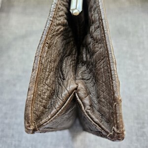 May include: A dark brown clutch with a textured, wrinkled surface and a silver-toned metal kiss-lock closure at the top. The bag features a gusseted side construction that allows it to expand, showcasing the deep brown material and stitching.