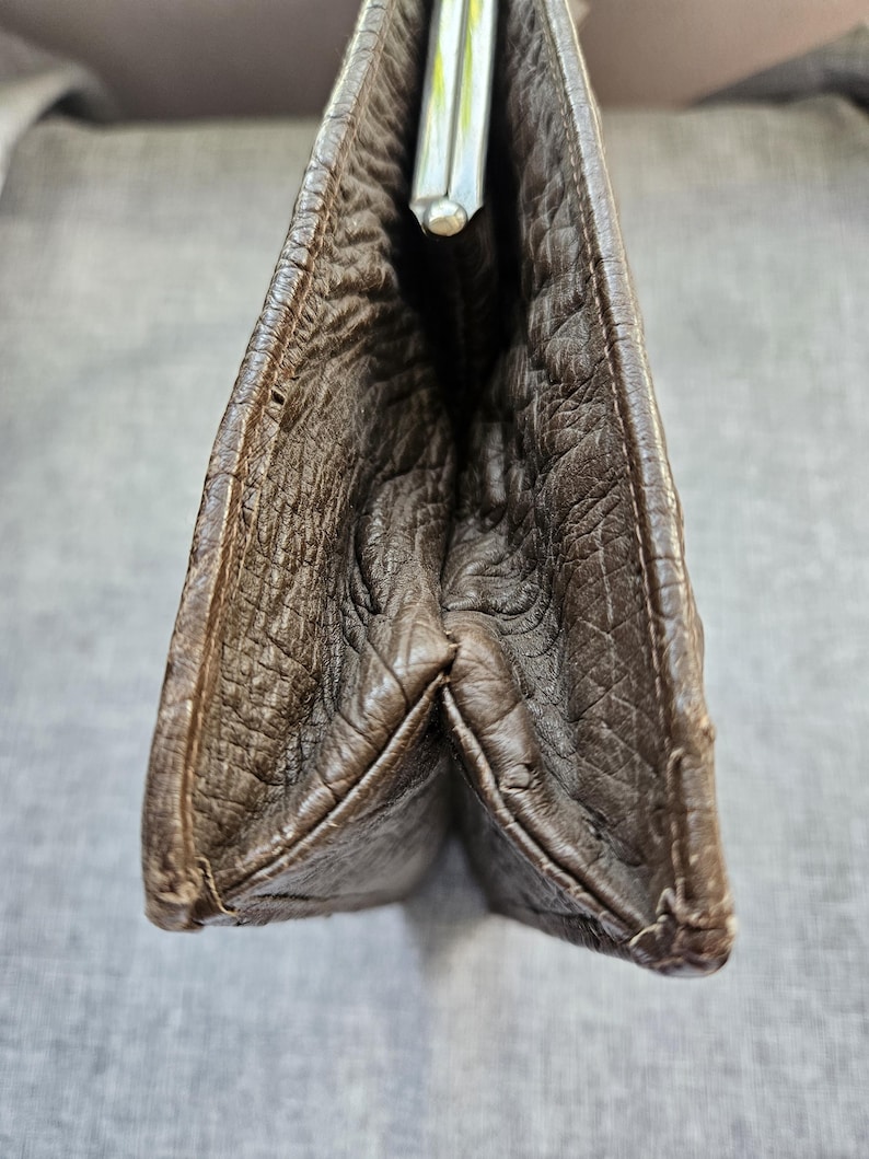 May include: A dark brown clutch with a textured, wrinkled surface and a silver-toned metal kiss-lock closure at the top. The bag features a gusseted side construction that allows it to expand, showcasing the deep brown material and stitching.