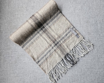 May include: A rectangular scarf featuring a plaid pattern in shades of beige, grey, and dark brown. The fabric has a soft, textured appearance with a folded top edge and a series of long, loose fringe tassels along the bottom edge.