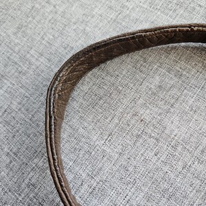 May include: A curved brown leather bag strap featuring a textured, pebbled surface with visible stitching along the edges. The strap displays signs of wear, including darkened areas and slight fraying along the seams, showing a worn-in appearance.