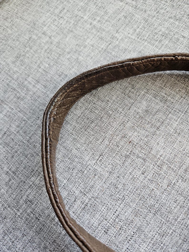 May include: A curved brown leather bag strap featuring a textured, pebbled surface with visible stitching along the edges. The strap displays signs of wear, including darkened areas and slight fraying along the seams, showing a worn-in appearance.