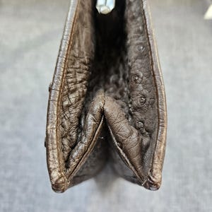 May include: A dark brown leather clutch with a distinct textured surface featuring small, raised quill-like bumps. The bag has a structured frame with a silver-toned metal clasp closure at the top, showing a gusseted side profile design.