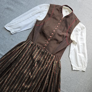 May include: A brown sleeveless bodice with thin red piping and a row of small buttons, worn over a white blouse with three-quarter length sleeves and lace detailing. The skirt features a dark brown, green, and red vertical striped pattern.