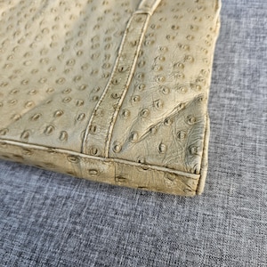 May include: A beige handbag featuring a textured surface with a repeating pattern of small, circular indentations resembling ostrich leather. The bag has a structured rectangular base with visible stitched seams along the edges and corners.