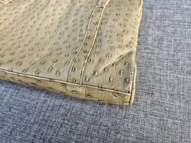 May include: A beige handbag featuring a textured surface with a repeating pattern of small, circular indentations resembling ostrich leather. The bag has a structured rectangular base with visible stitched seams along the edges and corners.