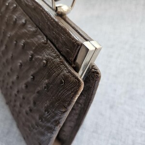 May include: A dark brown handbag with a textured ostrich-style finish featuring raised bumps. The bag has a structured rectangular frame with a silver-toned metal clasp closure at the top and a matching brown handle attached with silver hardware.