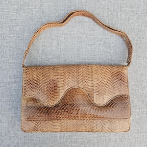 Pode incluir: Uma bolsa de ombro retangular com um padrão texturizado de pele de cobra castanha. A bolsa apresenta um design de aba ondulada distinto na parte frontal e uma alça de ombro única a condizer fixada nas laterais. O acabamento é castanho.