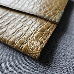 May include: Close-up of a tan, textured clutch bag with a crocodile skin pattern. The bag has a rectangular shape with a fold-over flap. The edges are finished with a metallic trim.