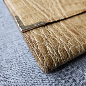 May include: A tan leather wallet with a crocodile skin texture. The wallet has a silver metal clasp and is folded closed. The leather has a worn appearance. The wallet is on a grey textured surface.