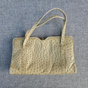 May include: A beige handbag with a textured ostrich-skin pattern, featuring a rectangular body, two thin shoulder straps, and a small gold-toned metal clasp closure at the top center. The bag has a structured design with vertical stitched detailing.