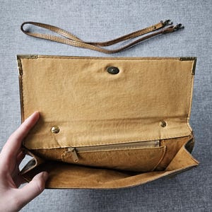 May include: Open, tan clutch bag with a matching shoulder strap. The interior has a zipped pocket and a snap closure. Gold-toned metal reinforces the corners. The bag is open, showing the inside.