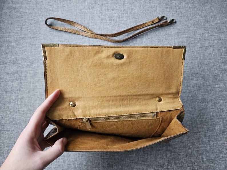 May include: Open, tan clutch bag with a matching shoulder strap. The interior has a zipped pocket and a snap closure. Gold-toned metal reinforces the corners. The bag is open, showing the inside.