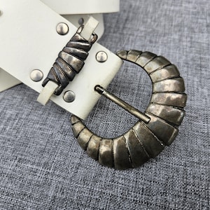 Puede incluir: Un cinturón blanco con una hebilla grande de metal plateado envejecido en forma de media luna con detalles acanalados. La correa está asegurada con remaches de metal plateado e incluye una placa metálica grabada con el texto von GERARD.