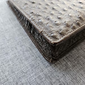 May include: A rectangular brown wallet featuring a distinct ostrich leather texture with raised quill bumps on the top surface and a darker, textured leather side panel with visible stitching along the edges of the folded construction.
