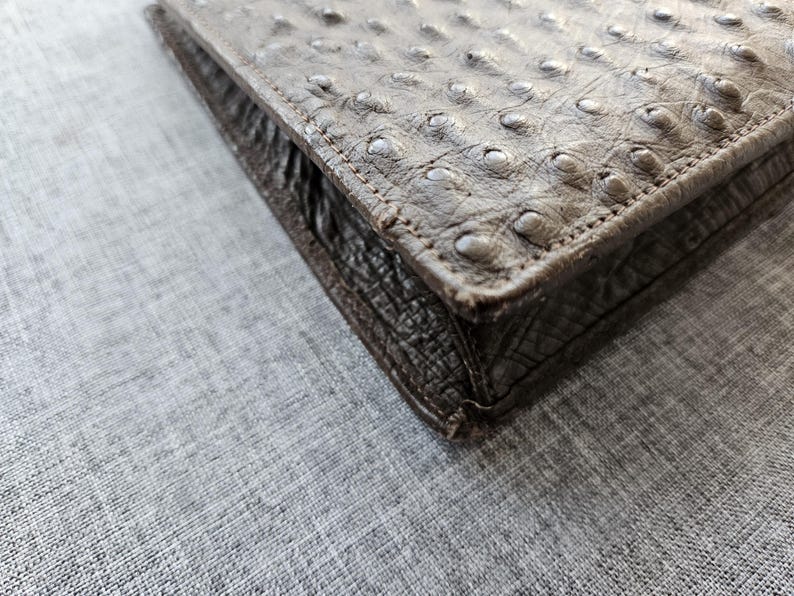 May include: A rectangular brown wallet featuring a distinct ostrich leather texture with raised quill bumps on the top surface and a darker, textured leather side panel with visible stitching along the edges of the folded construction.
