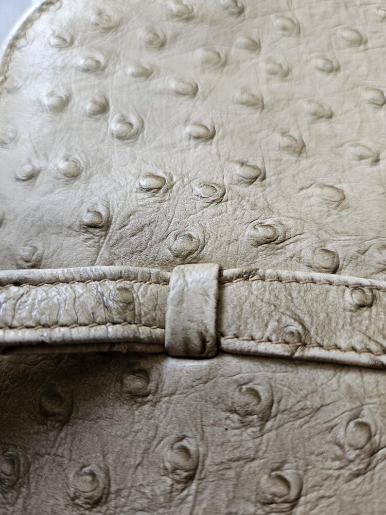 May include: A light beige textured material with a distinct quill pattern resembling ostrich leather. A horizontal strap with matching stitching and a central loop runs across the surface, creating a layered effect with the textured base.