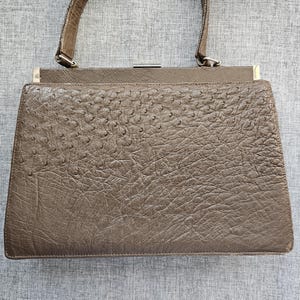 May include: A structured taupe handbag featuring a textured surface with a distinct quill pattern. The bag has a single top handle, a rectangular frame, and a metallic silver-toned clasp closure at the top center. The exterior displays a consistent, pebbled finish across the front panel.