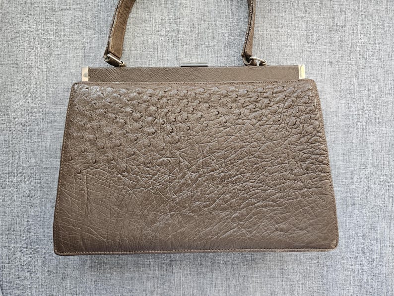 May include: A structured taupe handbag featuring a textured surface with a distinct quill pattern. The bag has a single top handle, a rectangular frame, and a metallic silver-toned clasp closure at the top center. The exterior displays a consistent, pebbled finish across the front panel.