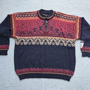 Könnte beinhalten: Ein dunkelblauer, langärmeliger Strickpullover mit Viertelreißverschluss und einem dekorativen Muster auf Brust und Schultern in Rot-, Creme- und Dunkelblautönen. Ein kleiner weißer Aufnäher mit schwarzem Text ist am linken Ärmel zu sehen.