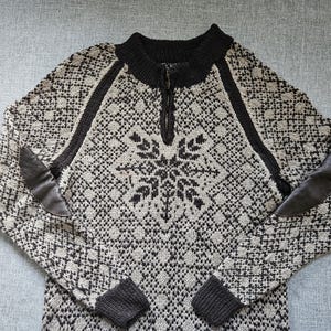 May include: A long-sleeve knit sweater featuring a black and beige geometric pattern with a central snowflake design. The sweater has a black ribbed mock neck with a short front zipper, black ribbed cuffs, and dark brown leather elbow patches.