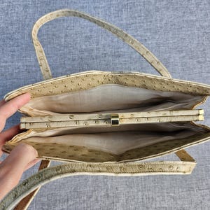 May include: A beige handbag with a textured ostrich-skin pattern, featuring a top-opening frame with a gold-tone metal clasp and a single shoulder strap. The interior is lined with a light-colored fabric and divided into two main compartments.