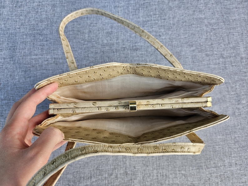 May include: A beige handbag with a textured ostrich-skin pattern, featuring a top-opening frame with a gold-tone metal clasp and a single shoulder strap. The interior is lined with a light-colored fabric and divided into two main compartments.