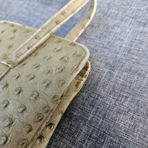 May include: A light beige handbag featuring a distinct textured pattern of raised circular quill follicles across the surface. The bag includes a thin, matching strap attached to the top edge, with visible stitching along the seams and edges.