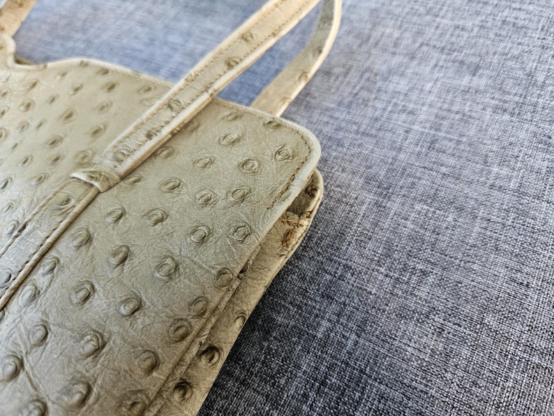 May include: A light beige handbag featuring a distinct textured pattern of raised circular quill follicles across the surface. The bag includes a thin, matching strap attached to the top edge, with visible stitching along the seams and edges.