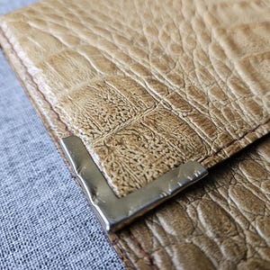 May include: Close-up of a tan leather wallet with a crocodile skin texture. The wallet has a silver metal corner piece and red stitching. The wallet is on a grey fabric surface.