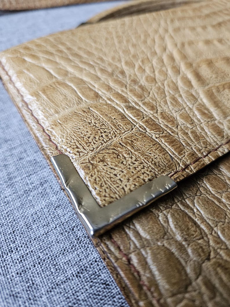 May include: Close-up of a tan leather wallet with a crocodile skin texture. The wallet has a silver metal corner piece and red stitching. The wallet is on a grey fabric surface.