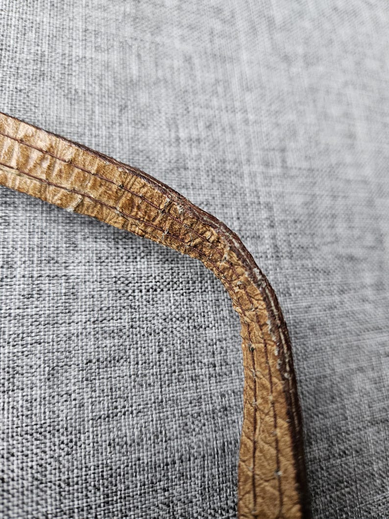 May include: Close-up of a section of a brown leather strap. The strap has a textured, patterned surface with visible stitching along the edges. The leather appears worn, with some areas showing signs of wear and tear.