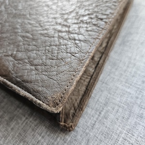 May include: A brown leather wallet featuring a distinct pebbled texture and visible stitching along the edges. The wallet has a folded construction with a thick, durable appearance and a matte finish, showcasing the natural grain of the material.