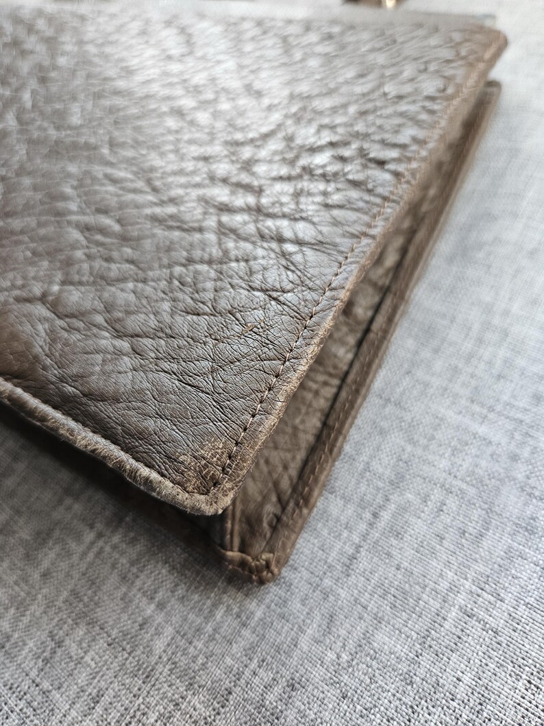 May include: A brown leather wallet featuring a distinct pebbled texture and visible stitching along the edges. The wallet has a folded construction with a thick, durable appearance and a matte finish, showcasing the natural grain of the material.