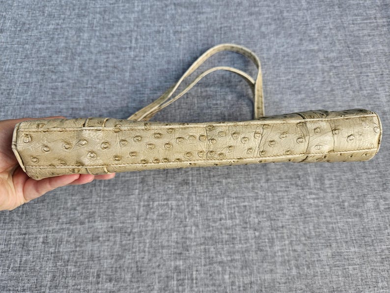 May include: A beige handbag featuring a textured ostrich-skin pattern with distinct quill follicles across the surface. The bag has a rectangular, slim profile and includes two thin, matching shoulder straps attached to the top edge.