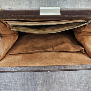 May include: A brown handbag with a textured exterior and a silver-toned metal frame. The interior is lined with tan fabric and features a zippered pocket along the top edge, a large open compartment, and a smaller slip pocket on the side.