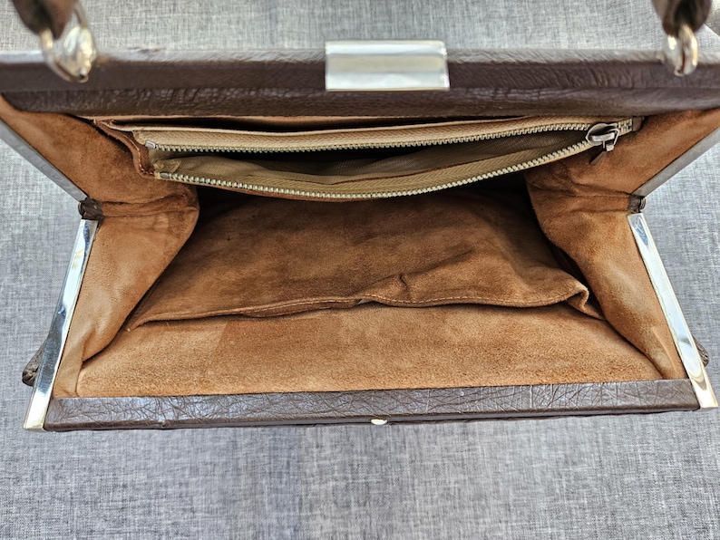May include: A brown handbag with a textured exterior and a silver-toned metal frame. The interior is lined with tan fabric and features a zippered pocket along the top edge, a large open compartment, and a smaller slip pocket on the side.