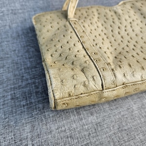 May include: A beige handbag featuring a distinct textured surface with raised quill follicles characteristic of ostrich leather. The bag includes a structured vertical panel design and a thin strap attachment visible at the top edge of the rectangular body.