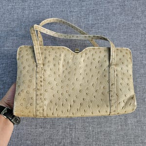 May include: A rectangular beige handbag featuring a textured ostrich leather pattern with distinct quill follicles. The bag has two thin top handles and a small gold-toned metal clasp closure at the center of the top edge, showcasing a structured design.