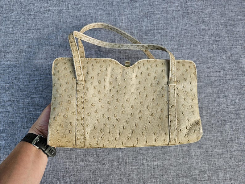 May include: A rectangular beige handbag featuring a textured ostrich leather pattern with distinct quill follicles. The bag has two thin top handles and a small gold-toned metal clasp closure at the center of the top edge, showcasing a structured design.