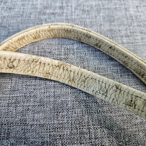 May include: Two off-white bag straps featuring a textured, cracked surface pattern with visible dark brown wear and aging along the edges and stitching. The straps are curved and display a distinct, weathered appearance with a worn-in finish.