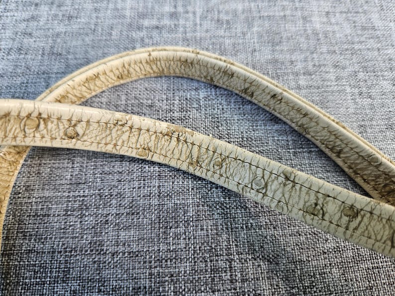 May include: Two off-white bag straps featuring a textured, cracked surface pattern with visible dark brown wear and aging along the edges and stitching. The straps are curved and display a distinct, weathered appearance with a worn-in finish.
