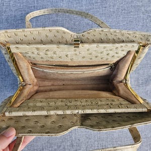 May include: A beige handbag with a textured ostrich-style pattern, featuring a gold-tone metal frame, a single top handle, and a light tan interior lining. The bag includes a zippered pocket inside and a secure metal clasp closure at the top.